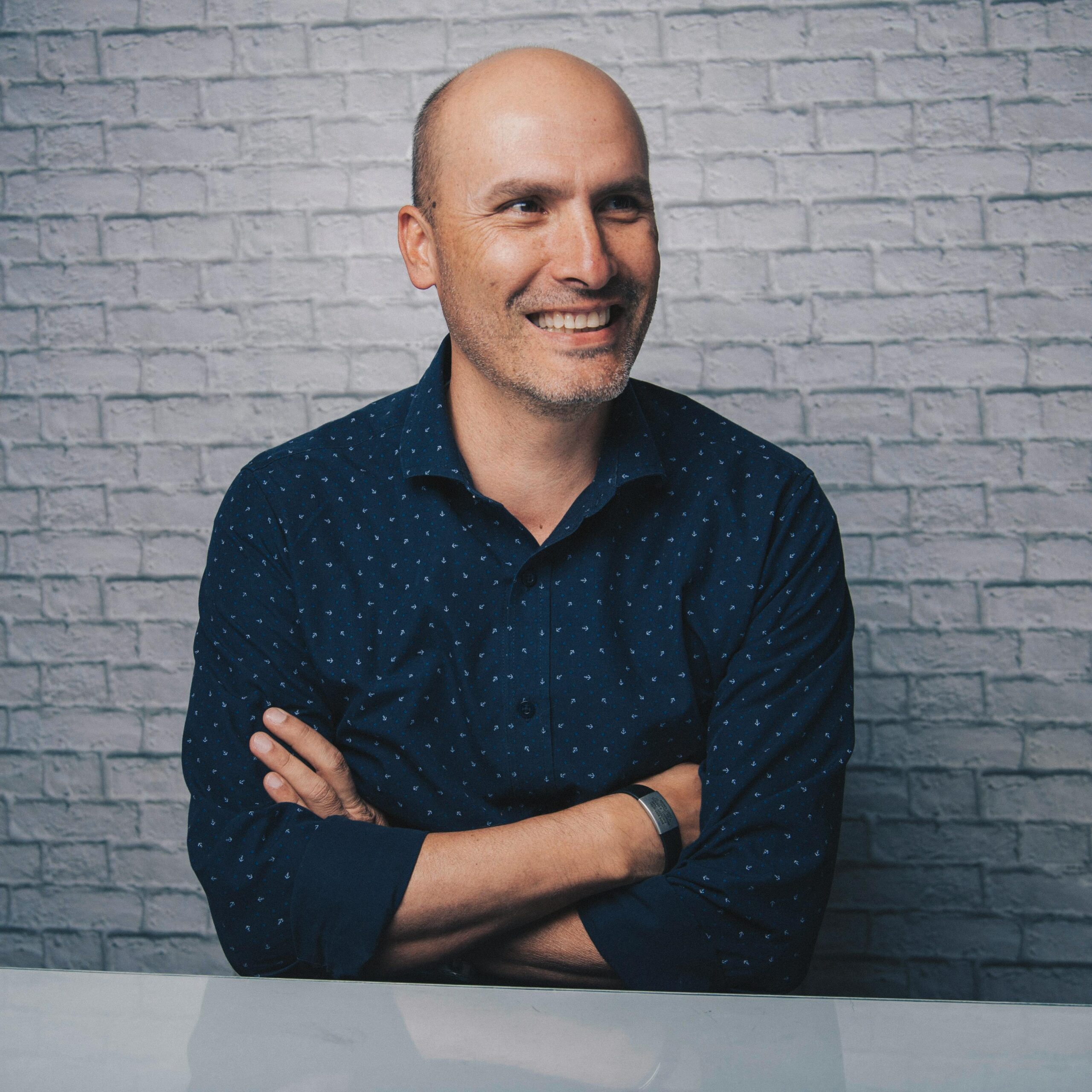 Positive bald male in stylish shirt sitting at white table with crossed arms while looking away and smiling on white brick wall